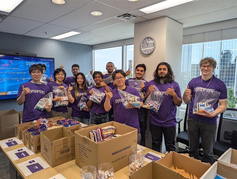 A group of interns in Synopsys shirts smiling and holding hygiene kits in an office space, surrounded by boxes of supplies and assembling kits.